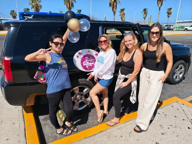 Bride and friends with Bride's Squad sign beside a black All Ways Suburban in Cabo